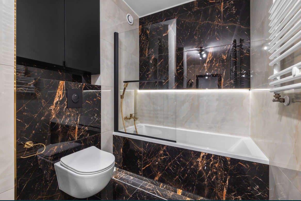 Elegancka łazienka z czarnymi marmurowymi płytkami z złotymi żyłami, podwieszana toaleta, złota armatura i podświetlana wanna za szklanym parawanem.