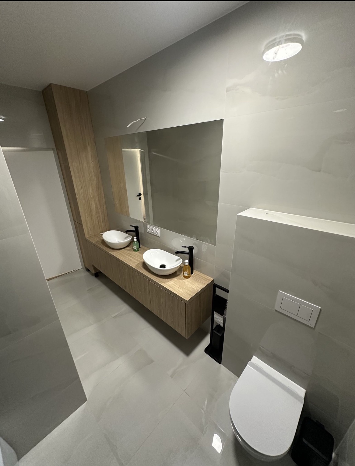 Nowoczesna łazienka z dwoma umywalkami nablatowymi, szafką z drewnopodobnym frontem i dużym lustrem, wykończona jasnymi płytkami imitującymi marmur, z widocznym czarnym stojakiem na papier toaletowy.