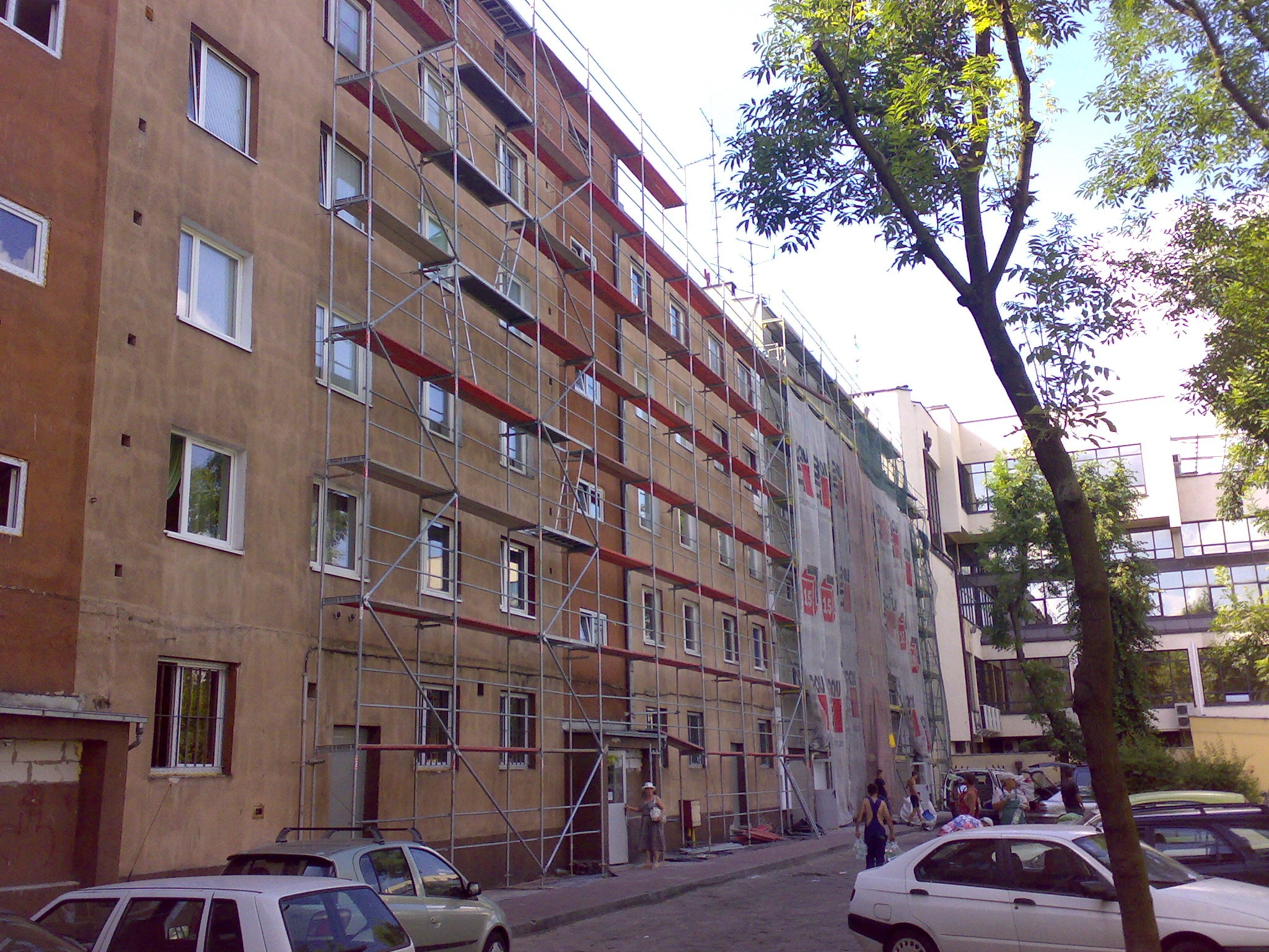 Budynek wielorodzinny w trakcie prac remontowych elewacji, widoczne rusztowania i siatka ochronna, na pierwszym planie zaparkowane samochody.