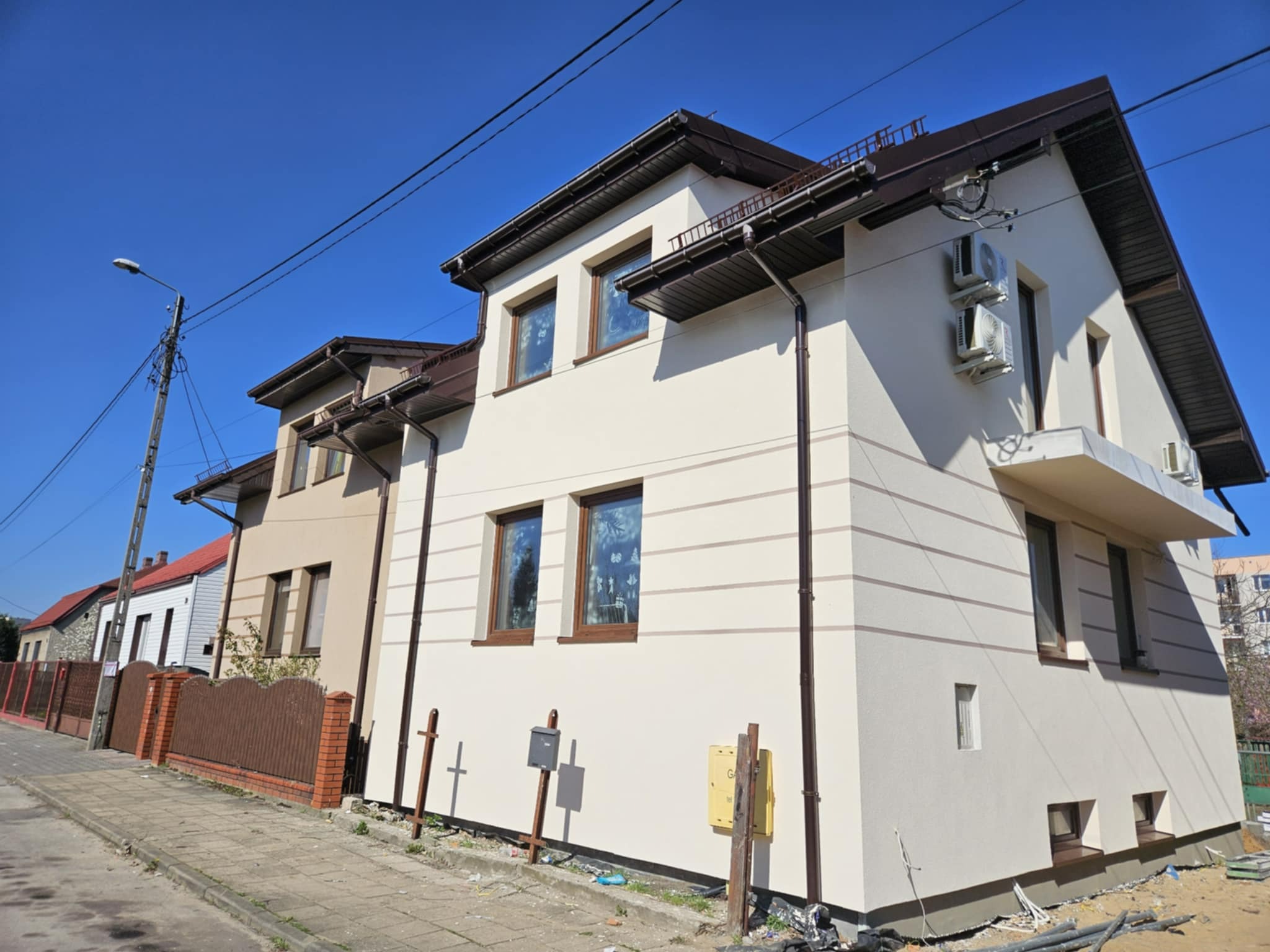 Dwupiętrowy dom z jasną elewacją, brązowymi ramami okiennymi i dwiema klimatyzacjami na ścianie. W tle widoczny fragment chodnika i sąsiednie budynki.