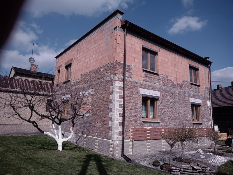 Dwukondygnacyjny dom w trakcie prac elewacyjnych, z fragmentami ocieplenia z czerwonej cegły i odsłoniętym murem ceglanym, widoczne okna z roletami, białe pnie drzew na pierwszym planie.