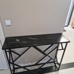 Firma Budowlano Kamieniarska Ireneusz Strzelczyk - Czarny blat z konglomeratu na metalowej konsoli o geometrycznym wzorze, ustawiony w nowoczesnym wnętrzu przy beżowej ścianie z widocznym włącznikiem światła.