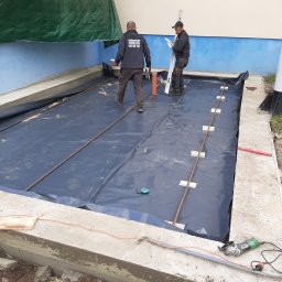 Firma Budowlano Kamieniarska Ireneusz Strzelczyk - Przygotowanie podłoża pod taras: rozłożona folia izolacyjna, zbrojenie z prętów stalowych stabilizowanych płytkami, dwaj pracownicy z poziomicą i narzędziami, widoczne krawędzie betonowej wylewki.