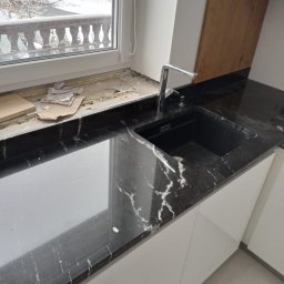 Firma Budowlano Kamieniarska Ireneusz Strzelczyk - Czarny, połyskujący blat z białym żyłkowaniem, zamontowany w kuchni, widoczny fragment okna z parapetem w trakcie prac wykończeniowych, bateria kuchenna ze stali nierdzewnej i czarny zlew.