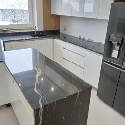 Firma Budowlano Kamieniarska Ireneusz Strzelczyk - Nowoczesna kuchnia z wyspą i czarnym blatem z wyraźnym, białym żyłkowaniem, białe szafki, okno z widokiem na zimowy krajobraz, czarna lodówka.