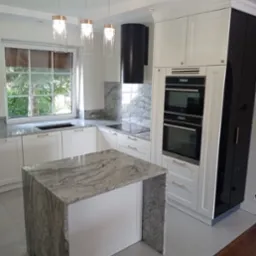 Jasna kuchnia z wyspą z szarego kamienia, białymi szafkami i czarnym okapem, oświetlona trzema wiszącymi lampami ze szklanymi kloszami.