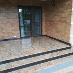 Firma Budowlano Kamieniarska Ireneusz Strzelczyk - Zadaszony taras z kamienną posadzką w odcieniach beżu i brązu, z trzema stopniami wykończonymi czarnym pasem, na tle ceglanej ściany budynku z brązowymi oknami.