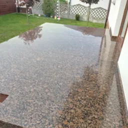 Błyszczący, brązowo-beżowy taras z kamienia naturalnego, odbijający korony drzew i siatkę bramki piłkarskiej w tle, przylegający do białej ściany budynku.