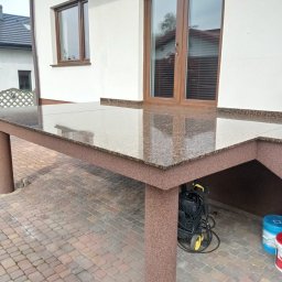 Firma Budowlano Kamieniarska Ireneusz Strzelczyk - Zewnętrzny stół z blatem z polerowanego kamienia w brązowych odcieniach, wsparty na solidnych, okrągłych nogach z tego samego materiału, ustawiony na brukowanym tarasie przed białym domem...