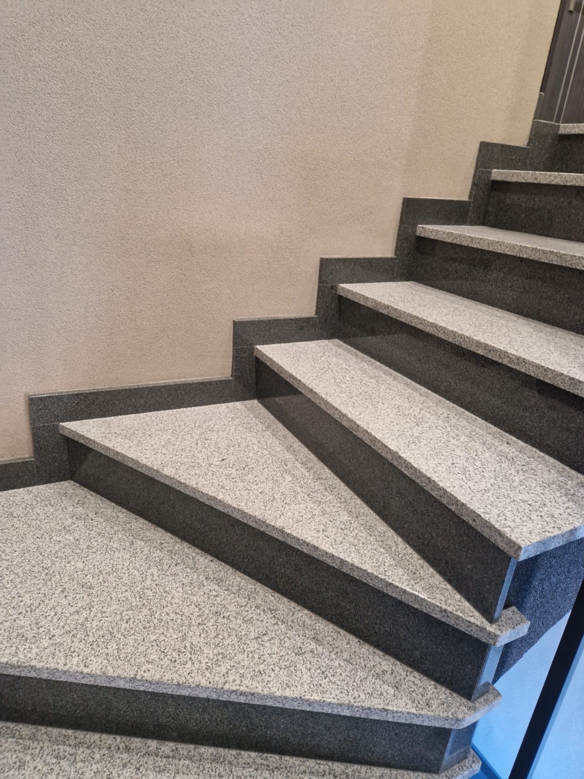 Schody wewnętrzne z szarego, nakrapianego kamienia z ciemnoszarymi krawędziami, widok z dołu na kilka stopni w górę, przy jasnej ścianie.