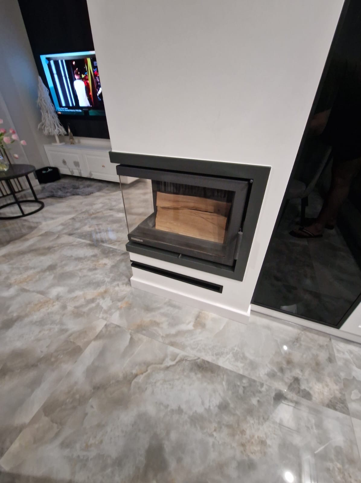 Nowoczesny kominek z przeszklonym frontem i polanami drewna wewnątrz, wbudowany w białą ścianę, na podłodze płytki imitujące marmur.