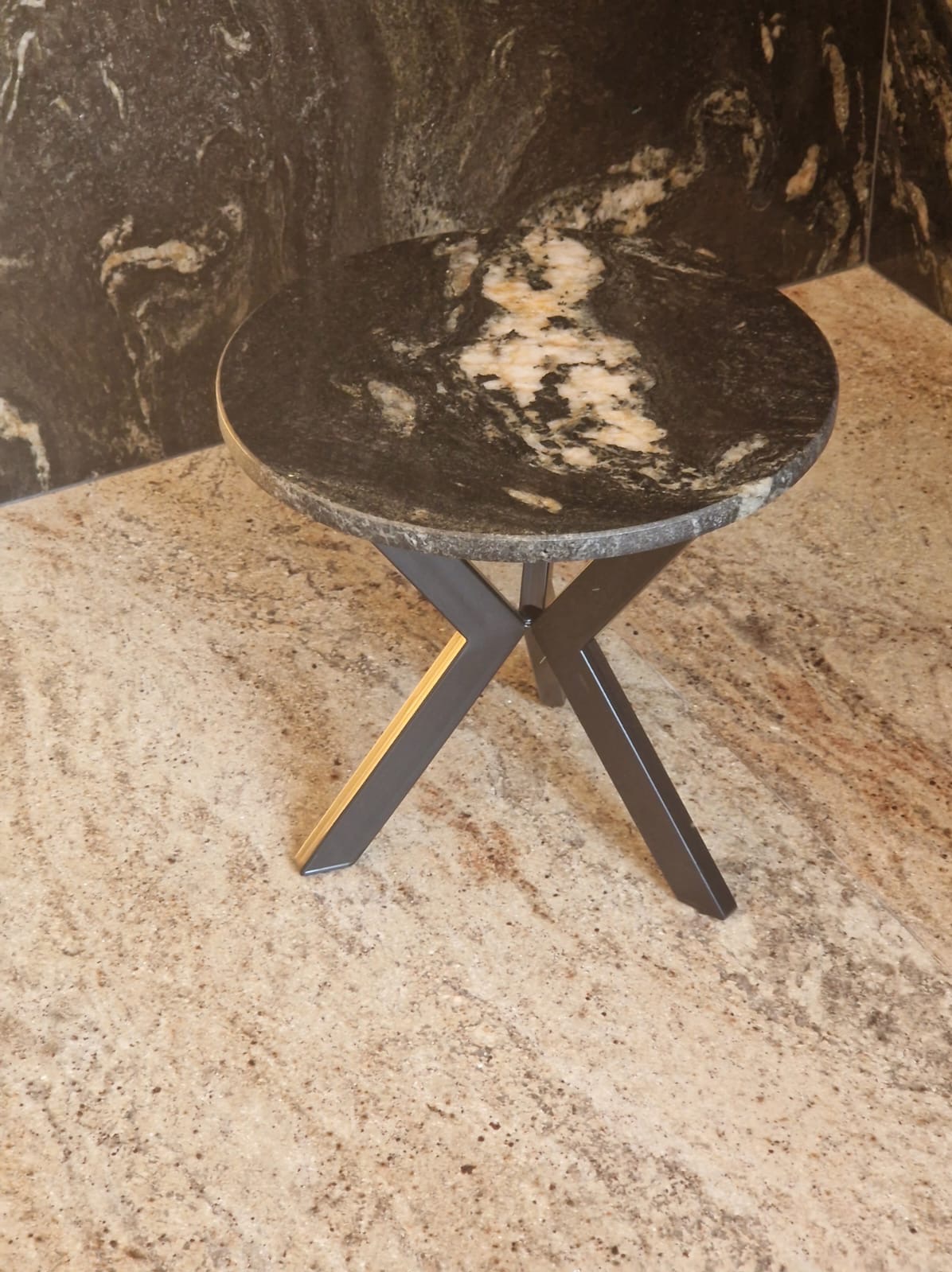 Okrągły stolik kawowy z blatem z ciemnego kamienia z jasnymi żyłkami, wsparty na metalowej podstawie z nogami w kształcie litery X, umieszczony na podłodze wyłożonej płytkami imitującymi kamień...