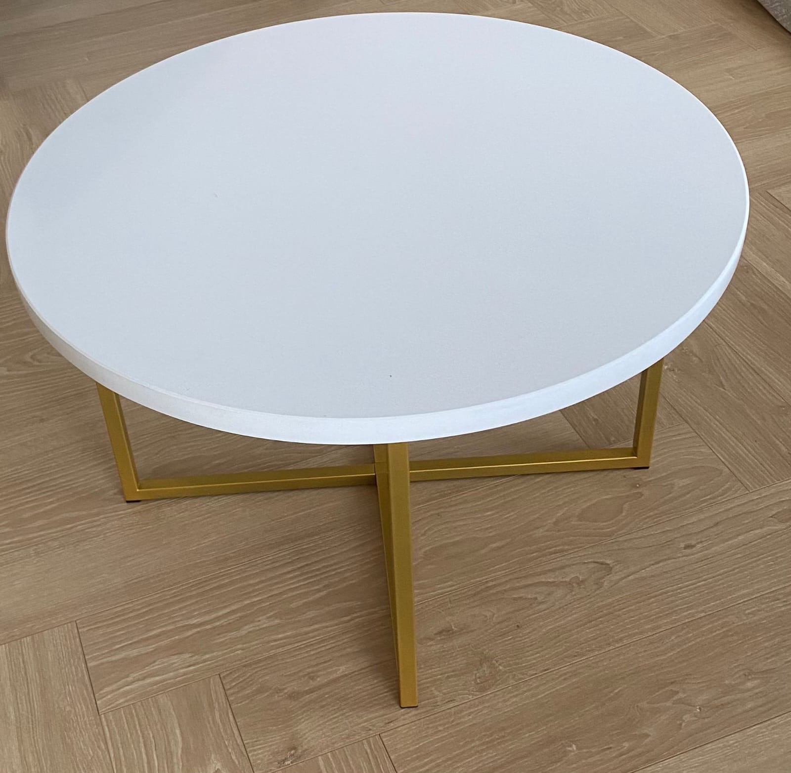 Okrągły stolik kawowy z białym blatem i złotymi, kwadratowymi nogami na podłodze z paneli imitujących drewno ułożonych w jodełkę.