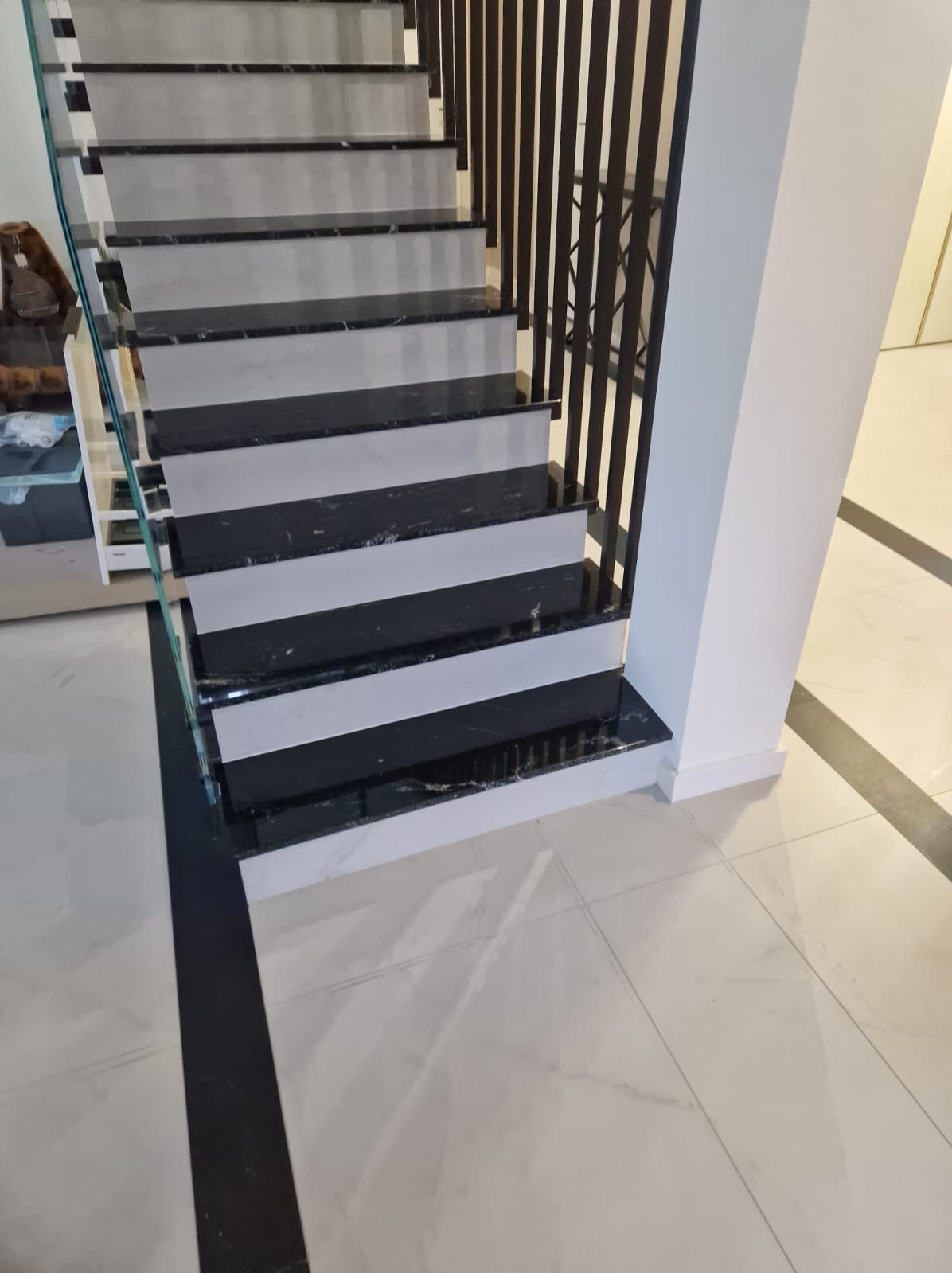 Nowoczesne schody wewnętrzne z czarnymi stopniami z kamienia i białymi podstopnicami, zabezpieczone szklaną balustradą i ciemną, pionową barierką, na jasnej marmurowej podłodze.