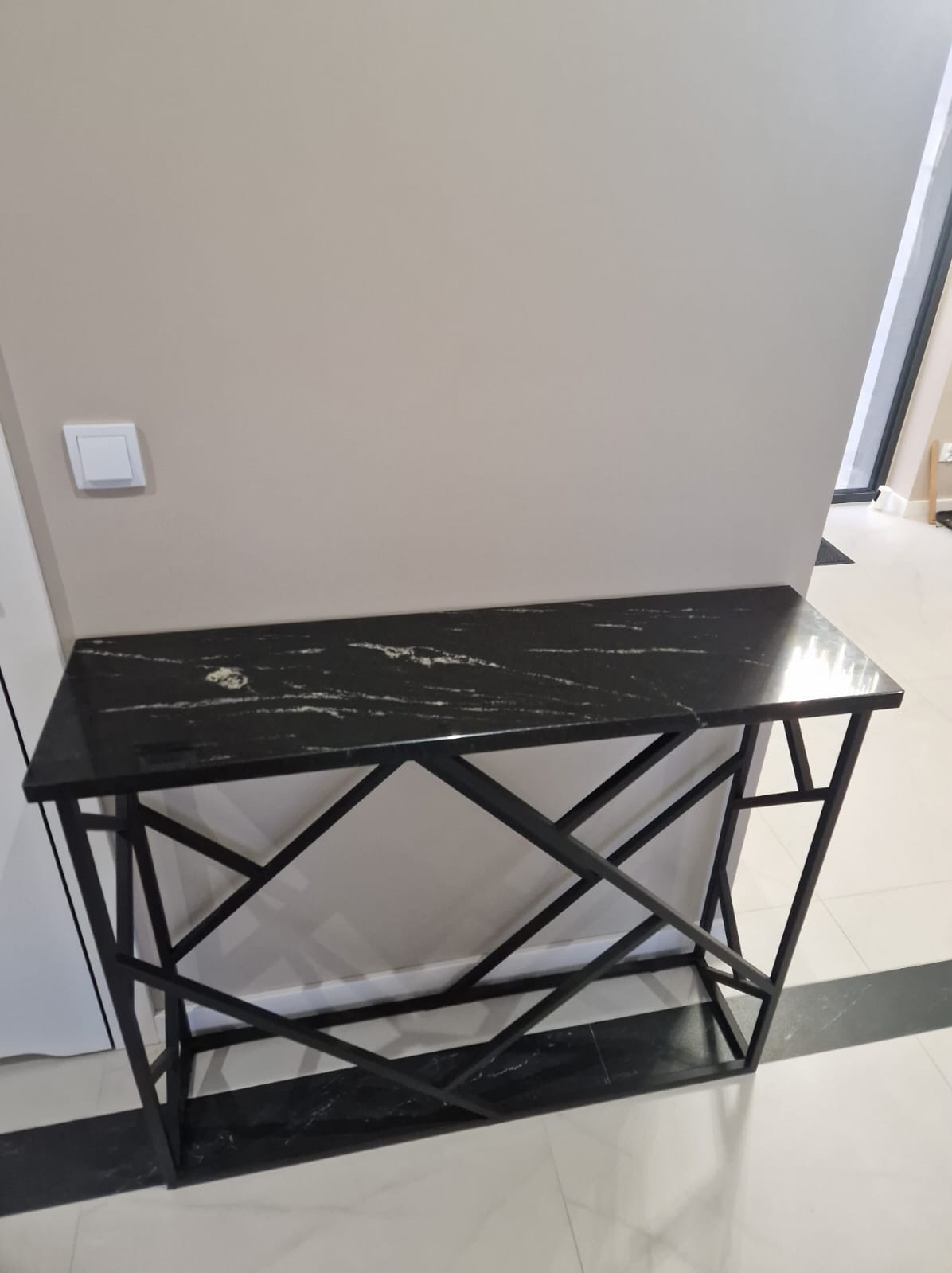 Czarny blat z konglomeratu na metalowej konsoli o geometrycznym wzorze, ustawiony w nowoczesnym wnętrzu przy beżowej ścianie z widocznym włącznikiem światła.