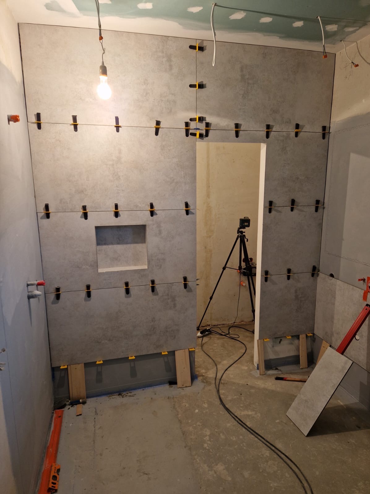 Montaż szarych płyt wielkoformatowych w łazience, widoczne systemy poziomowania płytek, otwór na półkę, przymocowane listwy przypodłogowe, surowy stan wykończenia podłogi i sufitu, statyw z laserem...