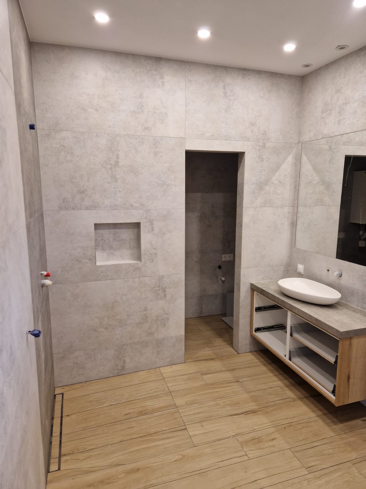 Nowoczesna łazienka z szarymi płytkami imitującymi beton, drewnopodobną podłogą, umywalką nablatową i wnęką w ścianie obok kabiny prysznicowej.