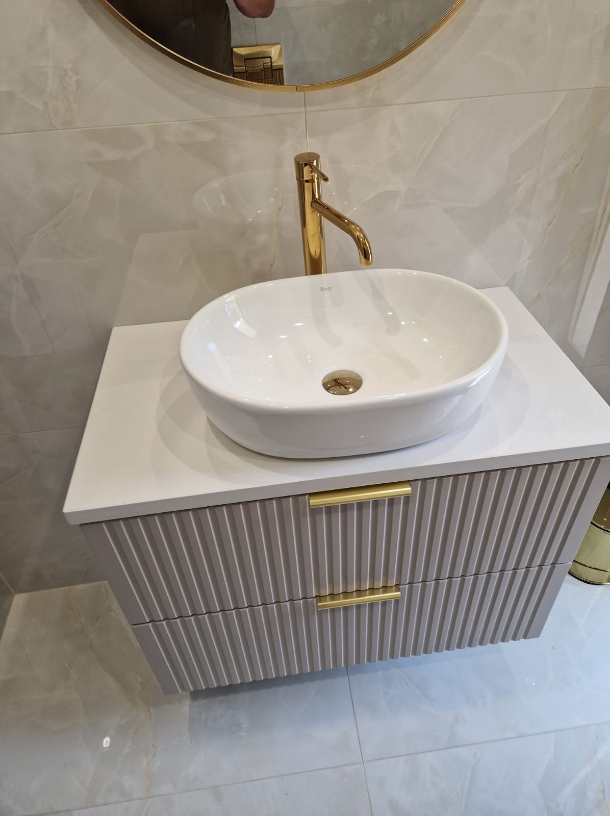 Nowoczesna łazienka z owalną umywalką nablatową, złotą baterią i szafką z frezowanymi frontami oraz blatem z konglomeratu, uzupełniona okrągłym lustrem w złotej ramie i płytkami imitującymi marmur.