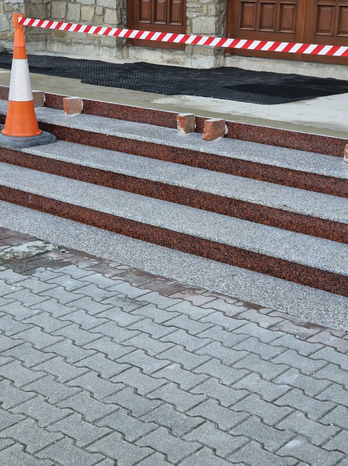 Granitowe schody zewnętrzne w trakcie montażu, zabezpieczone taśmą ostrzegawczą i pachołkiem, z widocznymi cegłami podtrzymującymi stopnie.