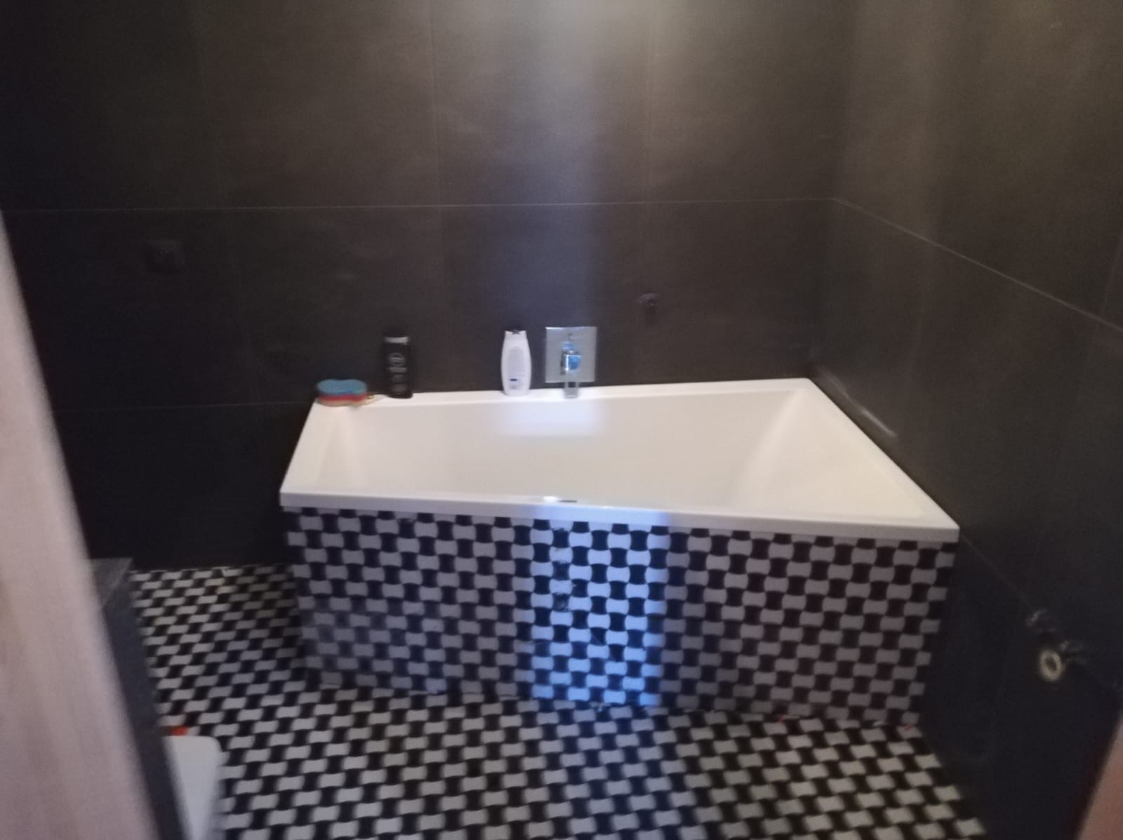 Nowoczesna łazienka z białą wanną obudowaną czarno-białą mozaiką, ciemne płytki na ścianach i podłodze w szachownicę, widoczne kosmetyki na brzegu wanny.