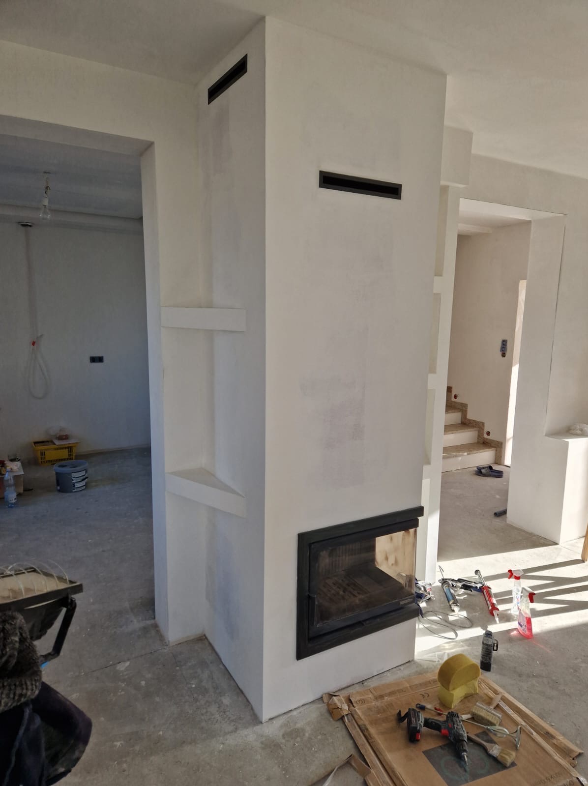 Wnętrze w trakcie wykańczania z nowoczesnym kominkiem z czarną obudową, widoczne narzędzia i materiały budowlane na podłodze, surowe ściany i przygotowane wnęki.