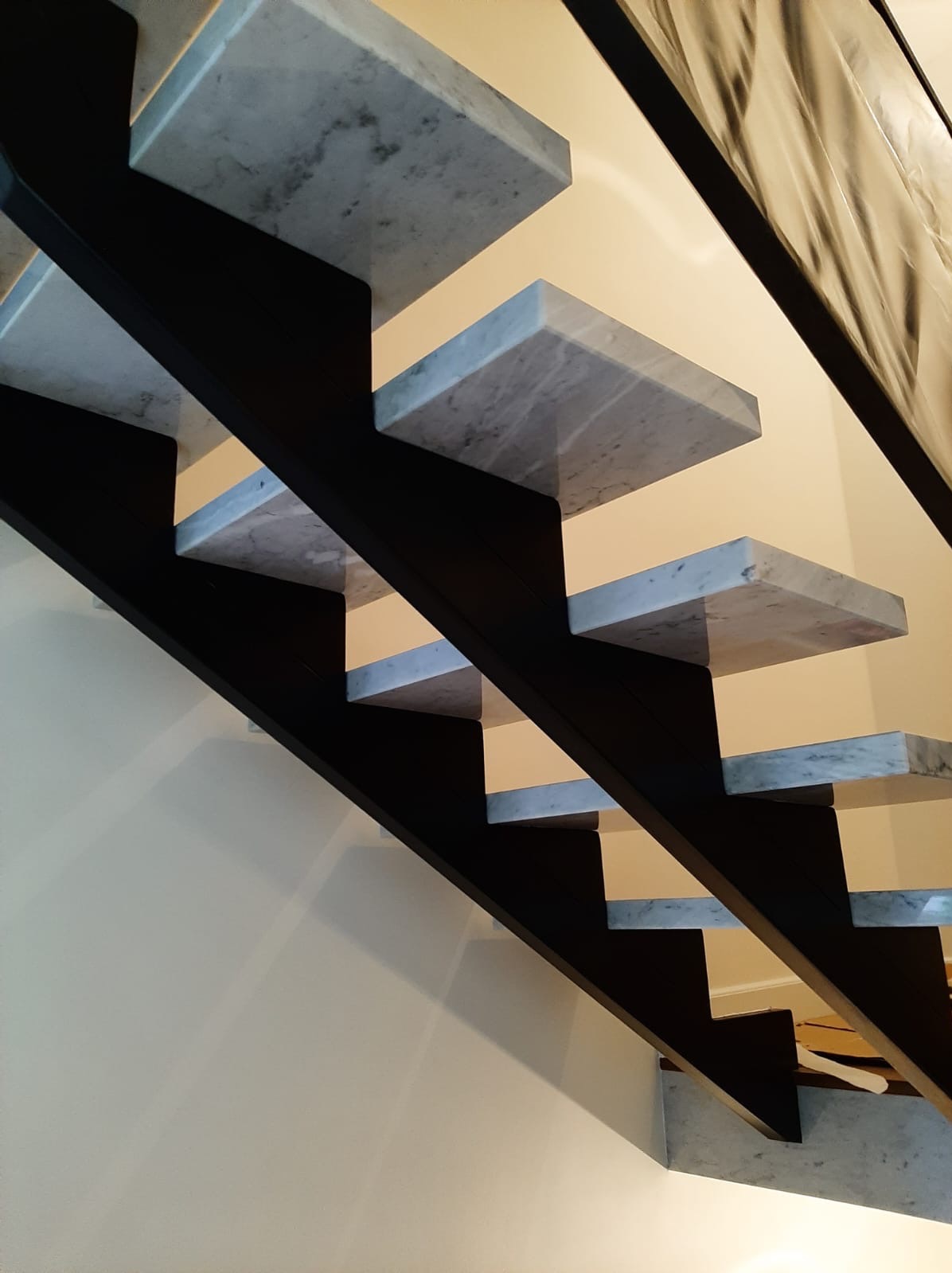 Nowoczesne schody wspornikowe z marmurowymi stopniami i czarną konstrukcją, widok z dołu na tle jasnej ściany.