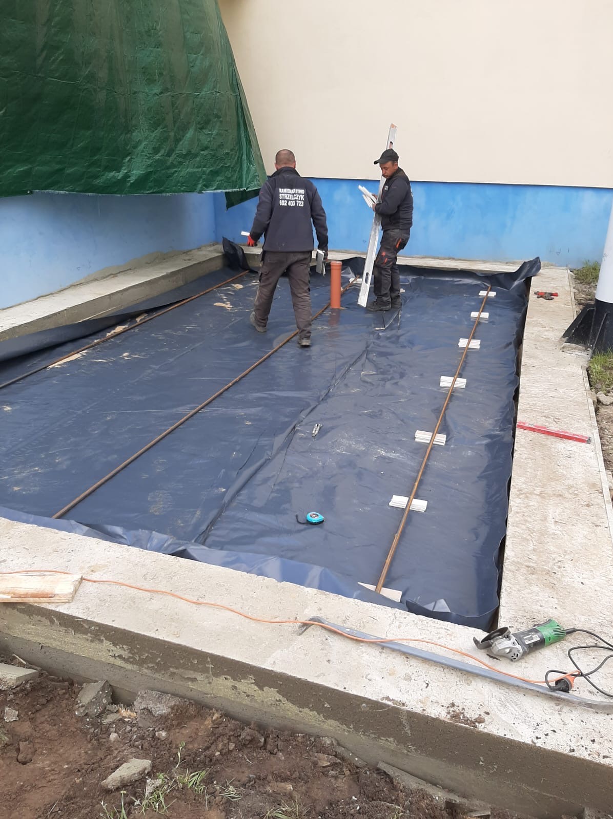 Przygotowanie podłoża pod taras: rozłożona folia izolacyjna, zbrojenie z prętów stalowych stabilizowanych płytkami, dwaj pracownicy z poziomicą i narzędziami, widoczne krawędzie betonowej wylewki.