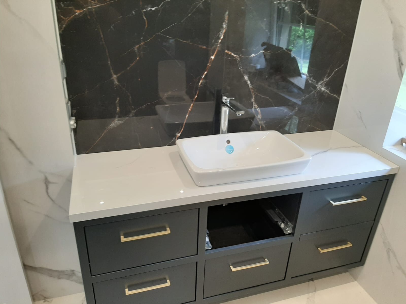Nowoczesna łazienka z umywalką nablatową, szarymi szafkami z złotymi uchwytami i blatem z białego konglomeratu, czarny panel ścienny z marmurowym wzorem, widoczne elementy instalacji wodnej.