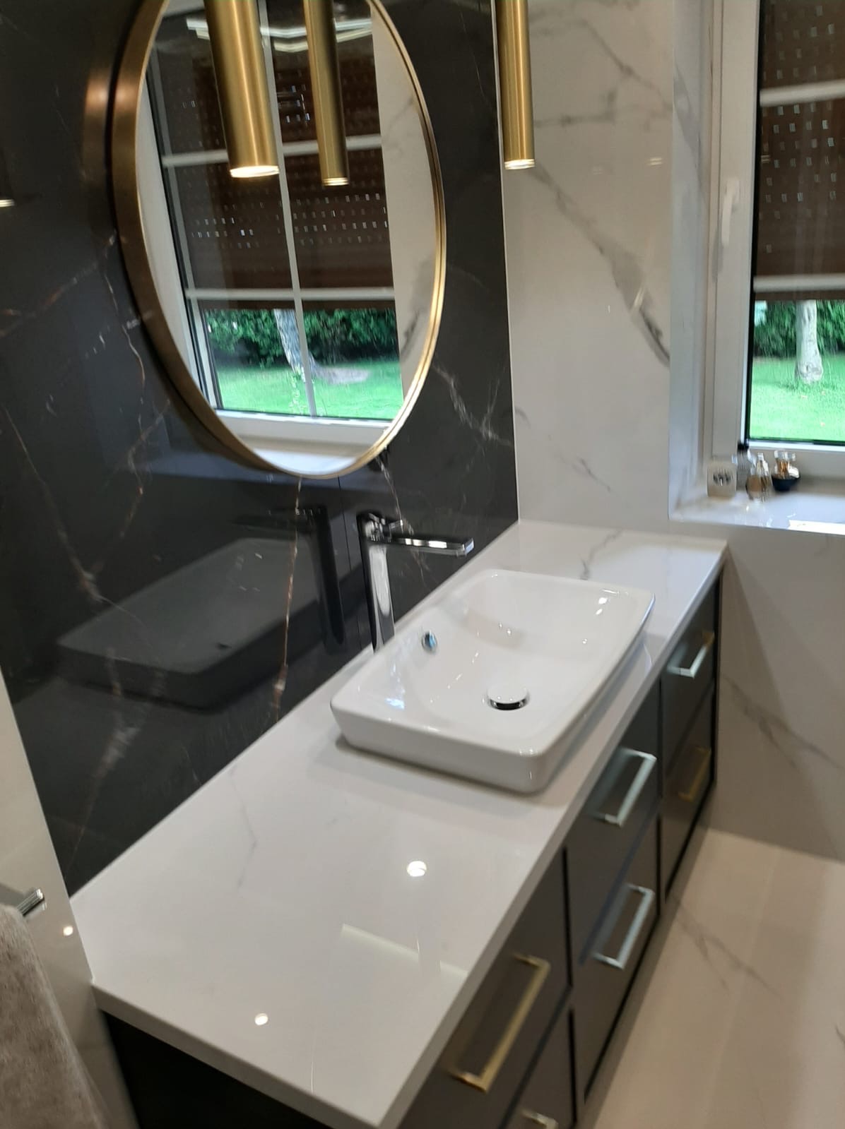 Nowoczesna łazienka z umywalką nablatową, blatem z białego konglomeratu, czarną ścianą z marmuru i okrągłym lustrem z oświetleniem wiszącym.