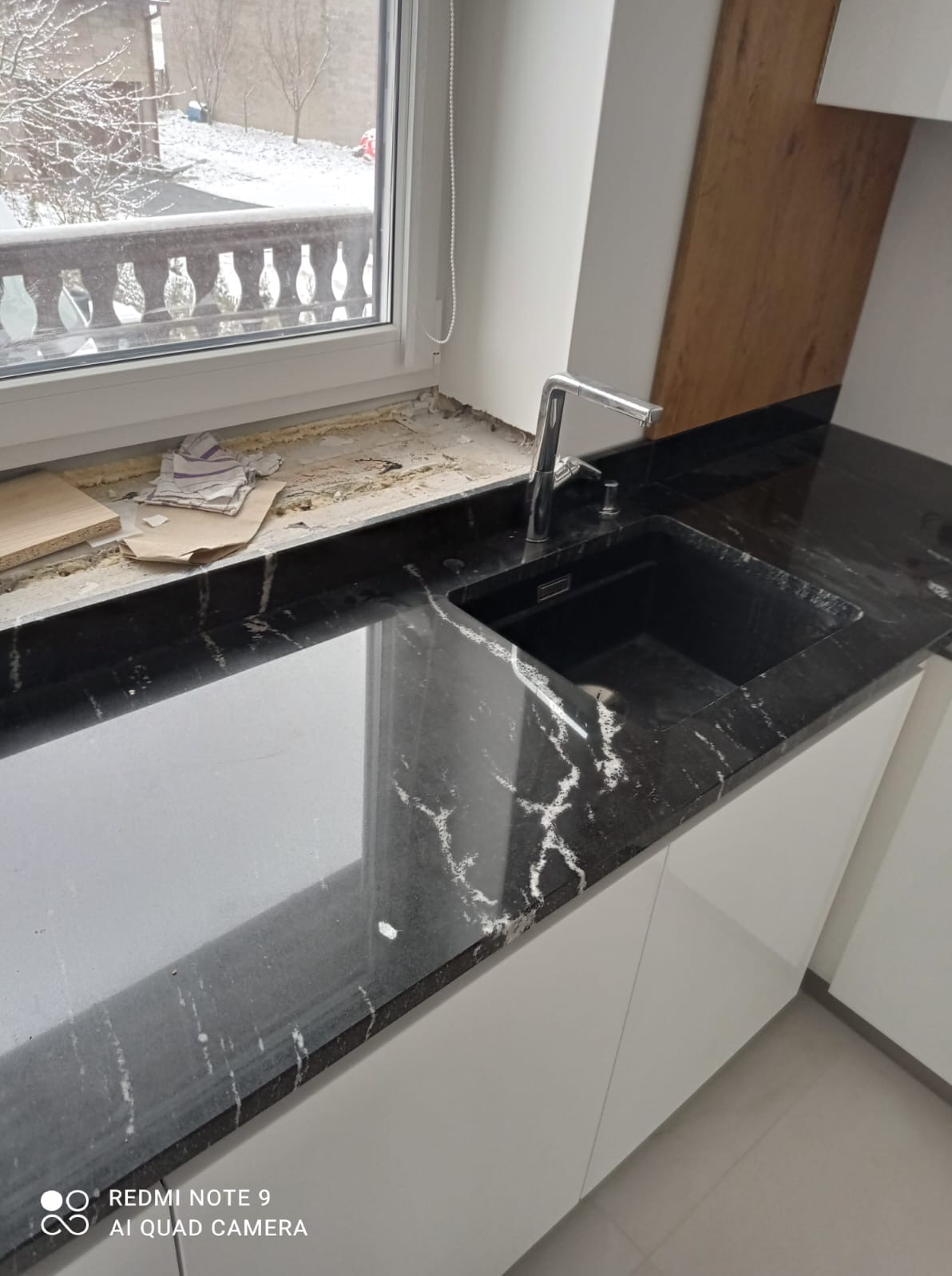 Czarny, połyskujący blat z białym żyłkowaniem, zamontowany w kuchni, widoczny fragment okna z parapetem w trakcie prac wykończeniowych, bateria kuchenna ze stali nierdzewnej i czarny zlew.
