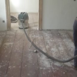 MADREM - Drewniana podłoga w trakcie renowacji. Widoczny szlifierka podłogowa z odkurzaczem i fragment osoby pracującej nad odnową parkietu.