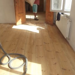 MADREM - Wnętrze z jasną, odnowioną podłogą z desek. W rogu odkurzacz przemysłowy. W tle osoba w drzwiach, wpadające światło słoneczne.