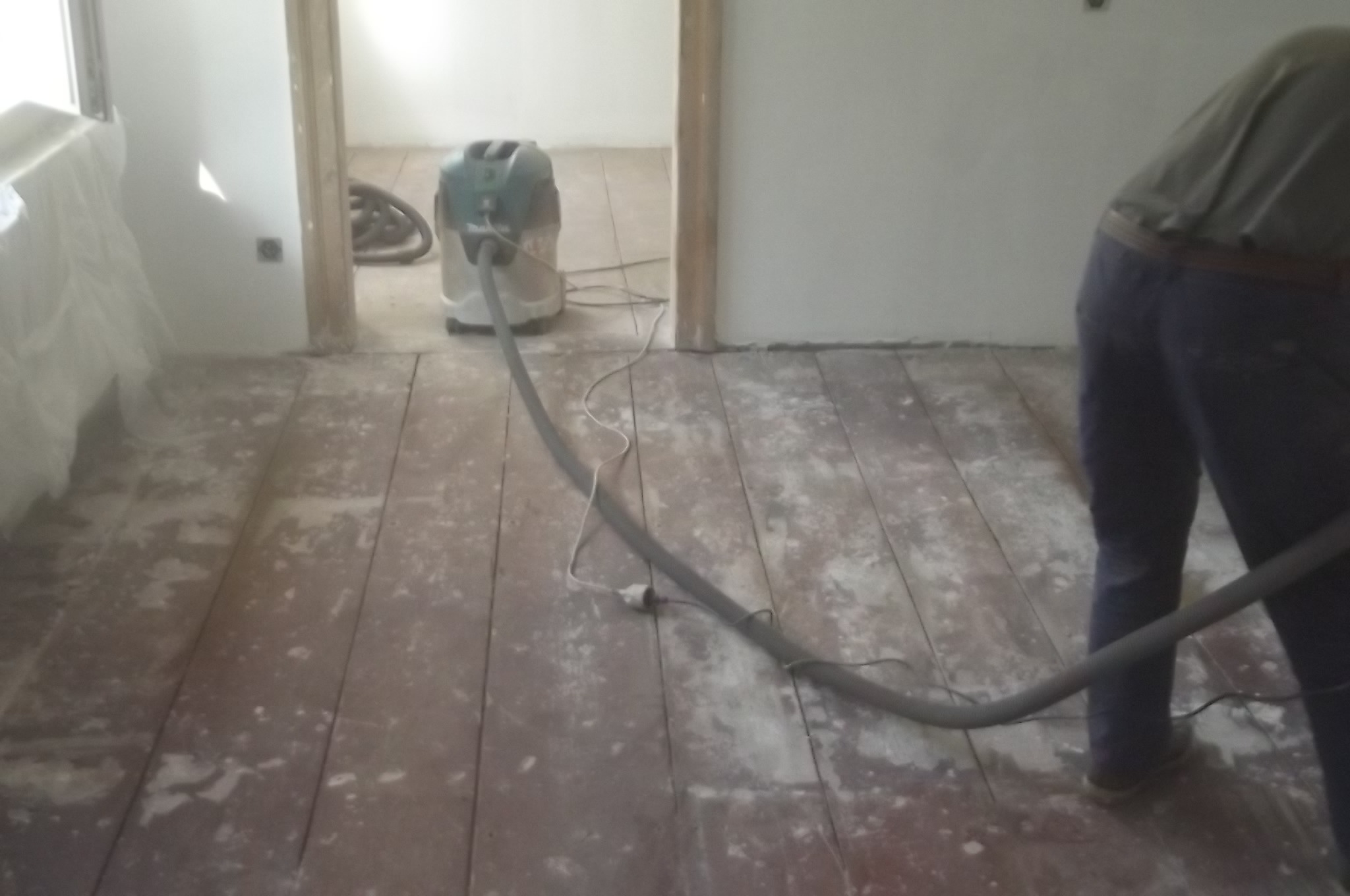 Drewniana podłoga w trakcie renowacji. Widoczny szlifierka podłogowa z odkurzaczem i fragment osoby pracującej nad odnową parkietu.