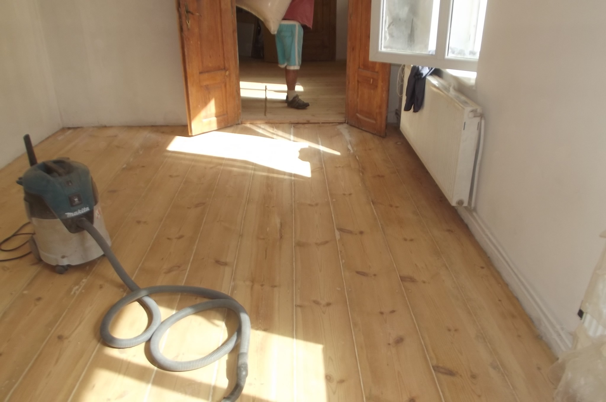 Wnętrze z jasną, odnowioną podłogą z desek. W rogu odkurzacz przemysłowy. W tle osoba w drzwiach, wpadające światło słoneczne.