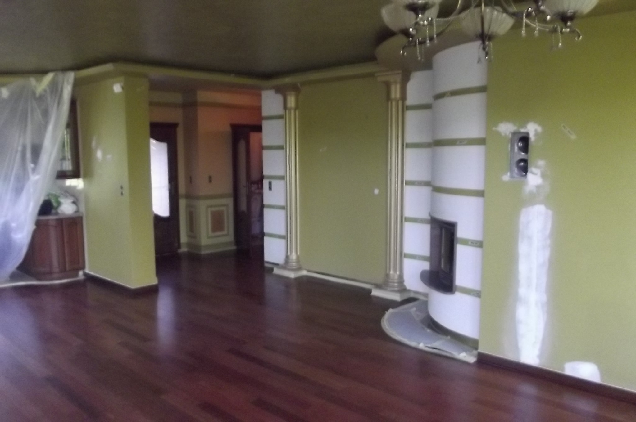 Salon z ciemną, lakierowaną podłogą, kolumnami i kominkiem w zabudowie. Ściany w odcieniach zieleni, częściowo przygotowane do malowania.