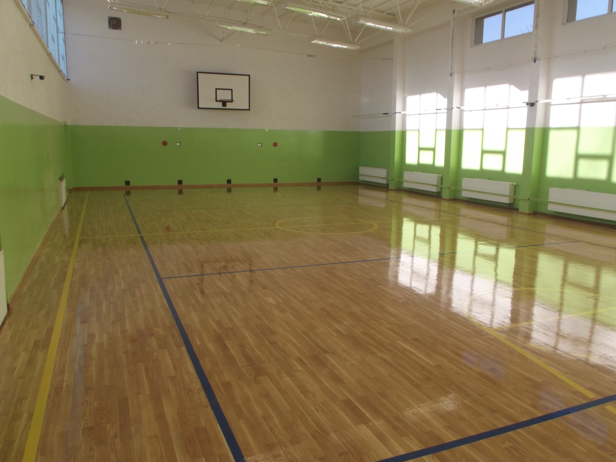 Błyszcząca, drewniana podłoga hali sportowej z liniami boiska, koszem do koszykówki na ścianie i oknami w tle, po renowacji.
