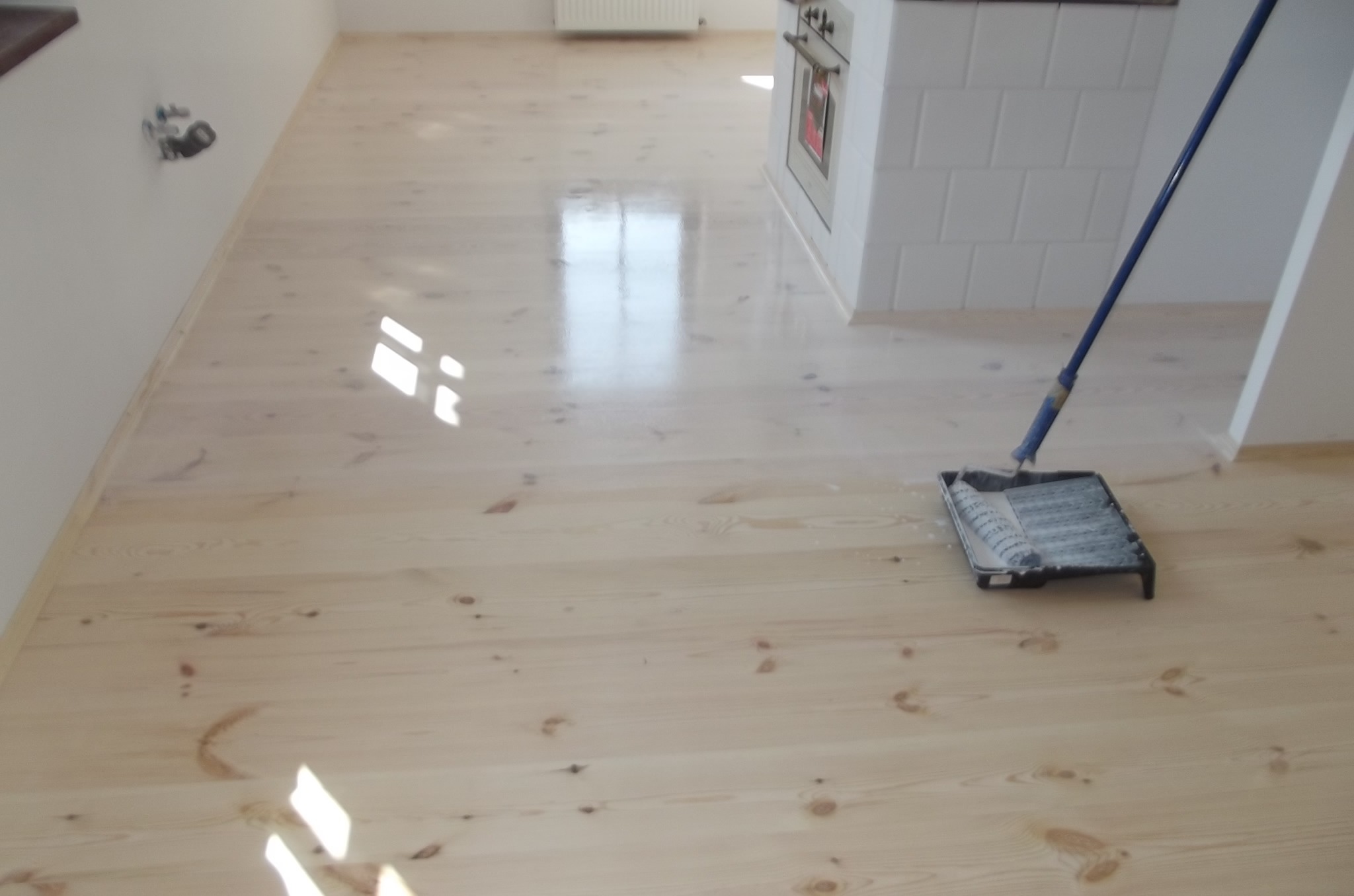 Jasna, odnowiona podłoga z desek po olejowaniu, widoczny wałek malarski i świeże refleksy światła, białe ściany i fragment zabudowy kuchennej w tle.