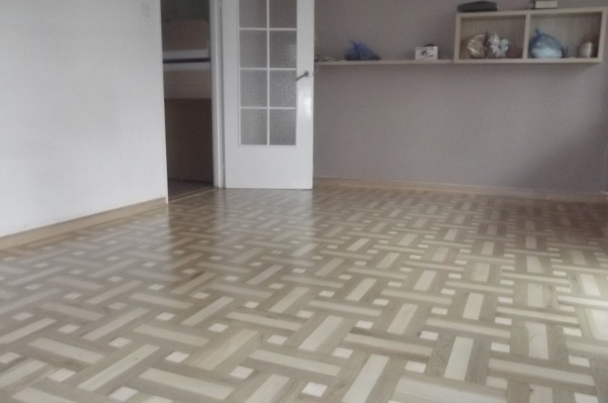 Parkiet w geometryczny wzór koszykowy, jasne drewno, widok na białe drzwi i półkę ścienną. Naturalne światło, minimalistyczne wnętrze.