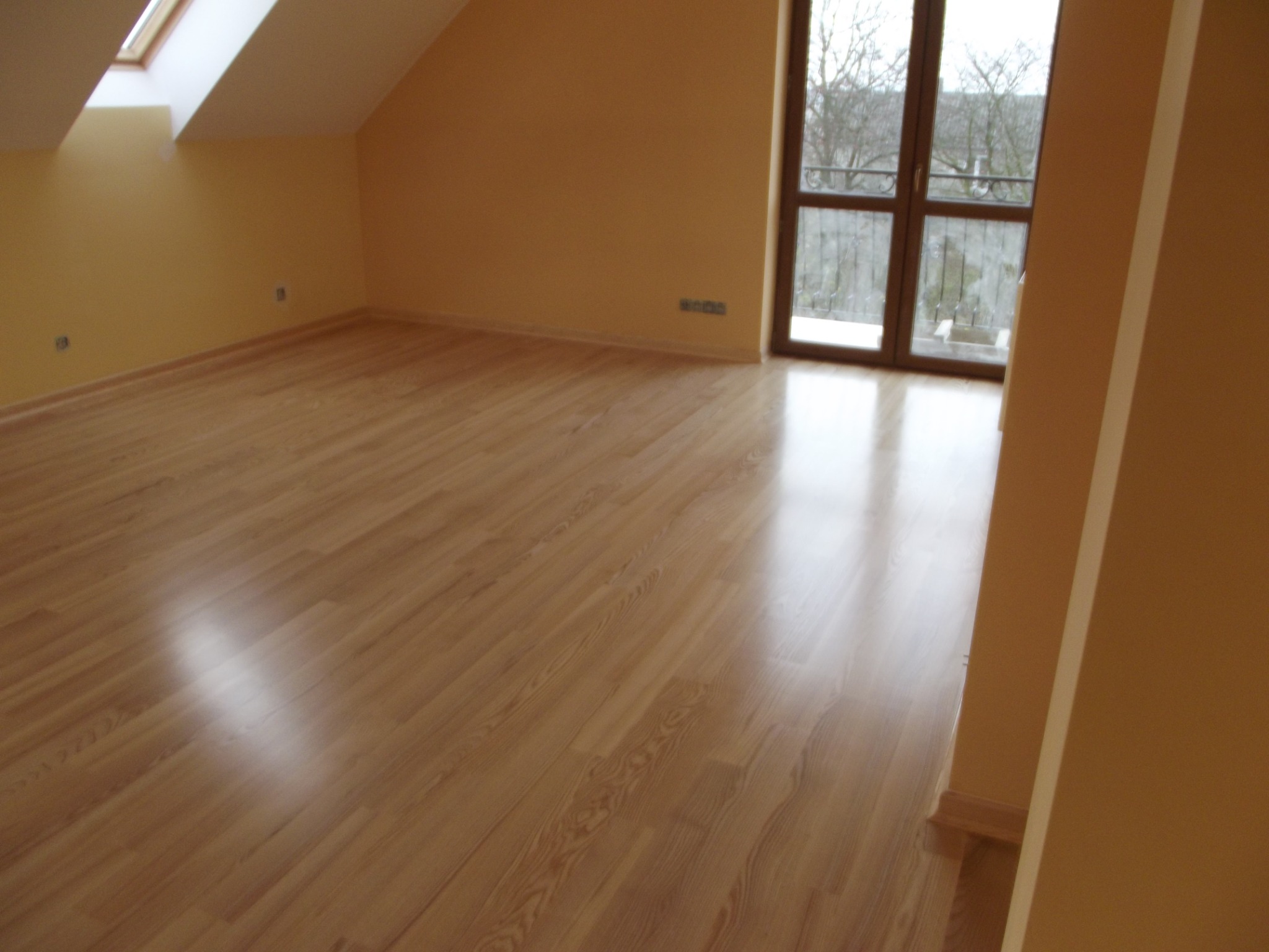 Jasny parkiet w wykończonym pokoju na poddaszu, widoczne okno balkonowe i skosy ścian. Świeżo położona podłoga w neutralnej kolorystyce.