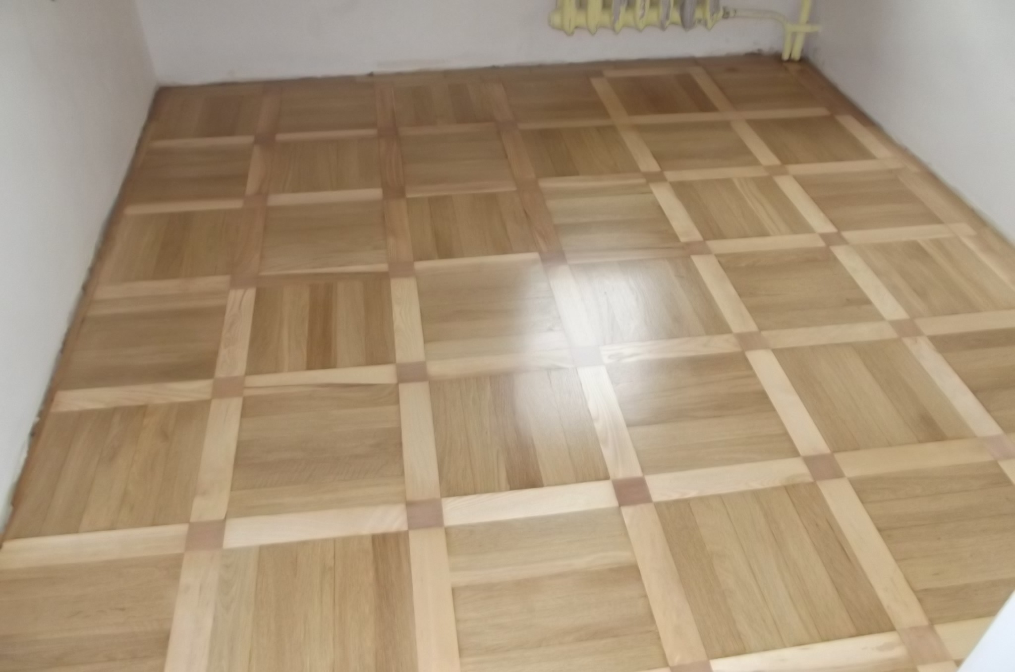 Jasny parkiet ułożony we wzór kwadratów z jaśniejszymi listwami, widoczny fragment grzejnika przy suficie, świeżo po renowacji.