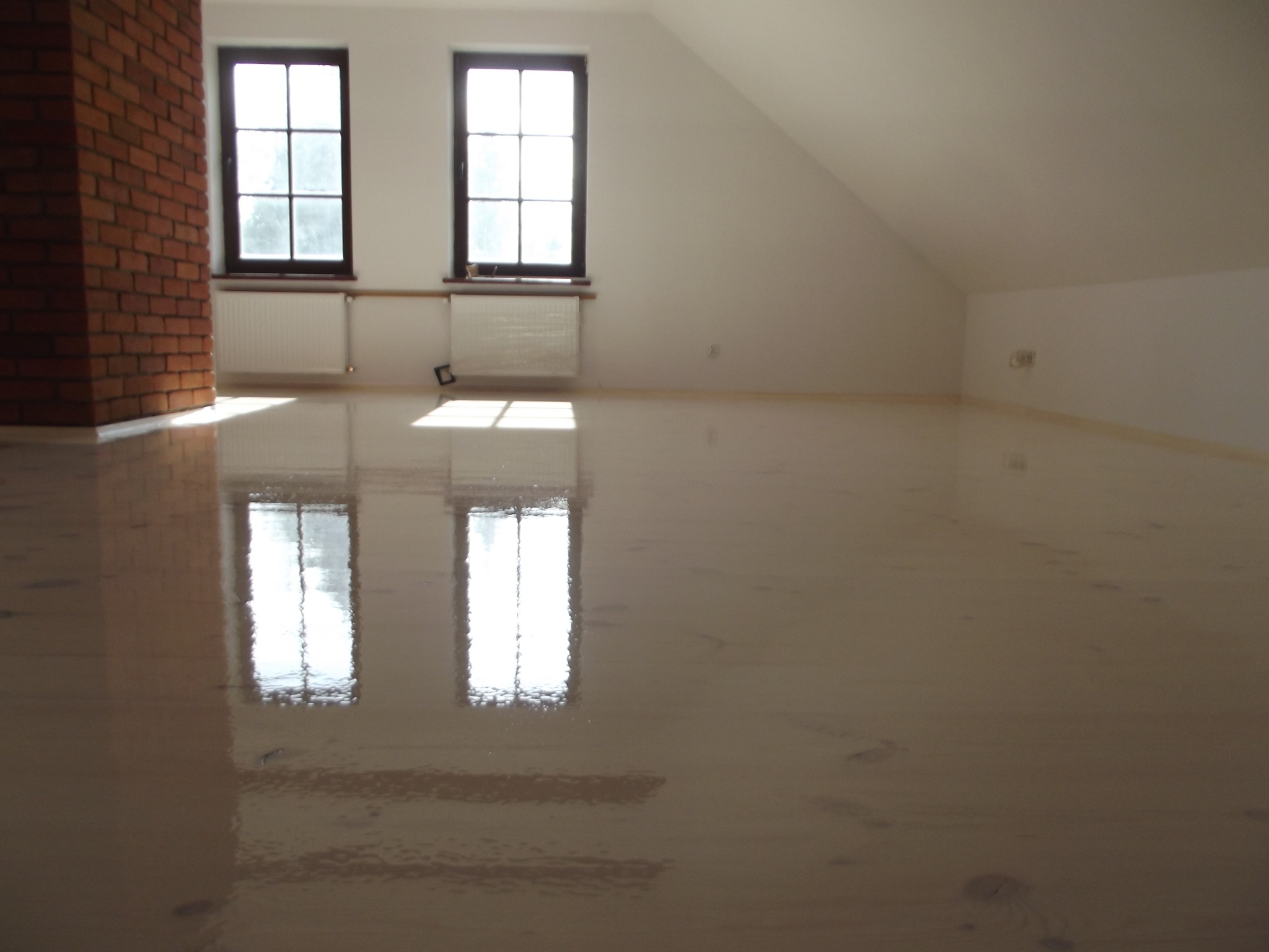 Błyszcząca, jasna drewniana podłoga w pokoju na poddaszu z ceglanym kominem i oknami. Odbicie okien w podłodze. Widoczne ślady po cyklinowaniu.