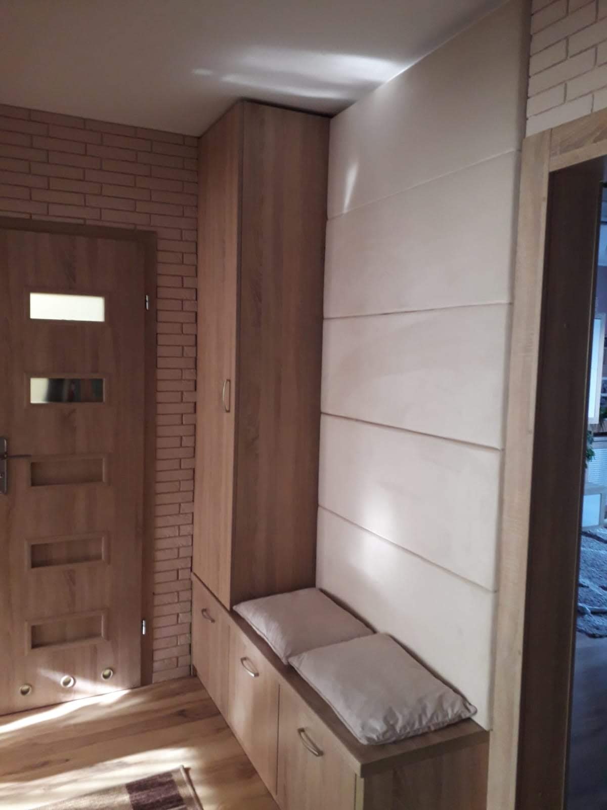 Wnęka w przedpokoju z zabudową z szafą i siedziskiem z poduszkami, obok drzwi z ceglaną ścianą, całość w odcieniach brązu i beżu.