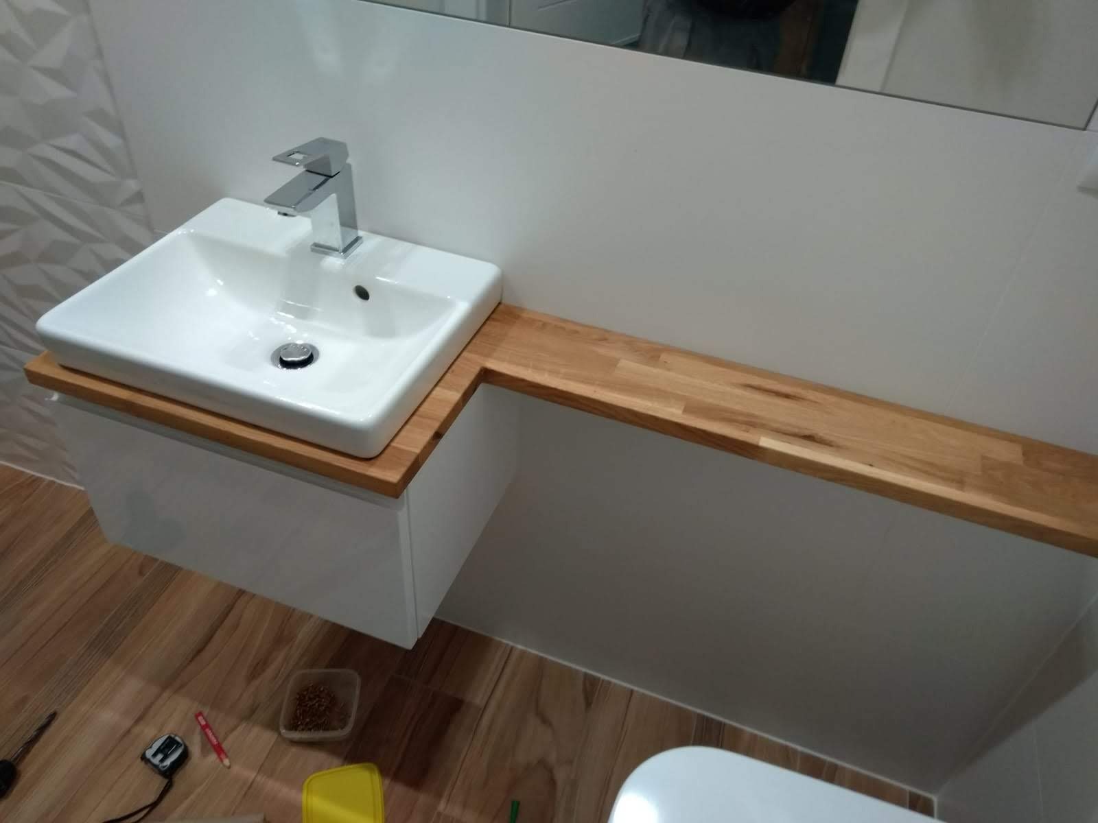 Biała umywalka nablatowa z chromowaną baterią, osadzona na białej szafce z blatem z jasnego drewna, widoczne narzędzia i akcesoria montażowe na podłodze.