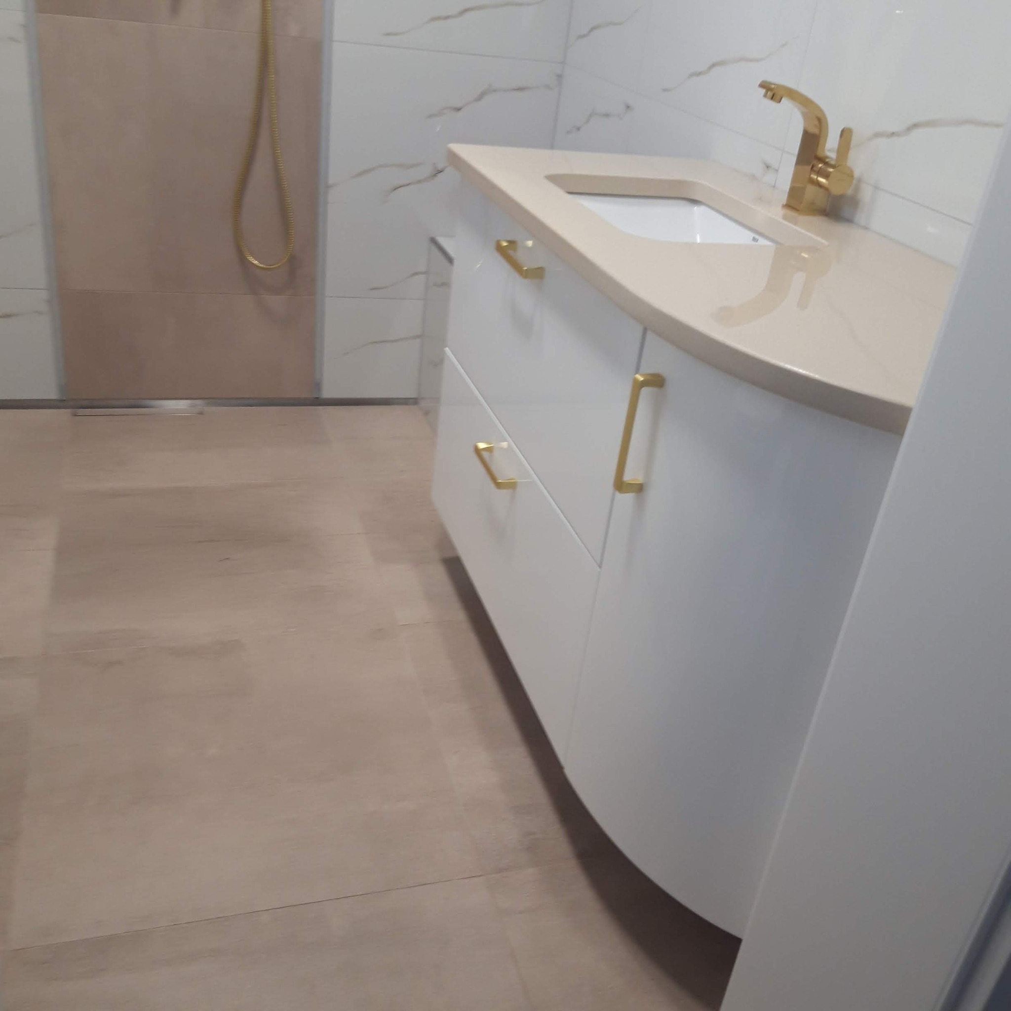 Biała szafka łazienkowa z beżowym blatem i złotą armaturą, widoczna na tle beżowo-białych płytek w łazience.