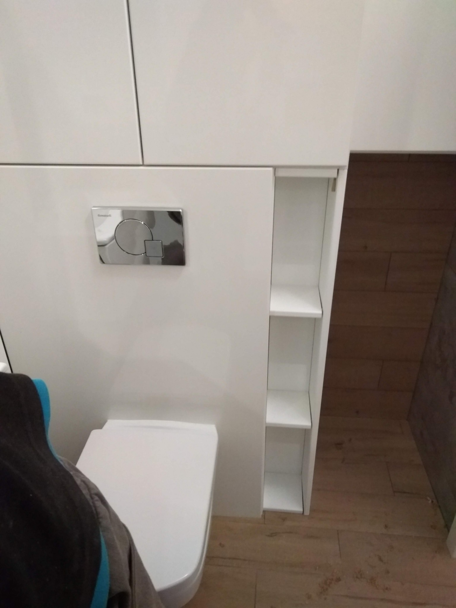 Biała zabudowa toalety z wnęką z półkami, chromowany przycisk spłukiwania, widoczna deska sedesowa i fragment drewnopodobnej podłogi.