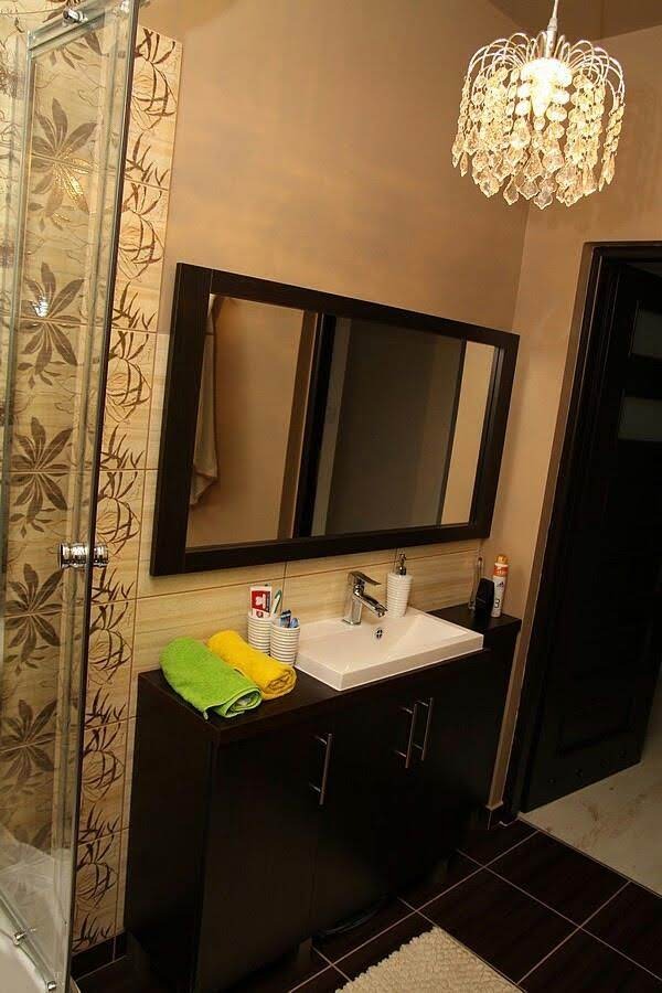 Nowoczesna łazienka z ciemnobrązową szafką pod umywalką, prostokątnym lustrem w ciemnej ramie, kabiną prysznicową z wzorzystymi płytkami i kryształowym żyrandolem.