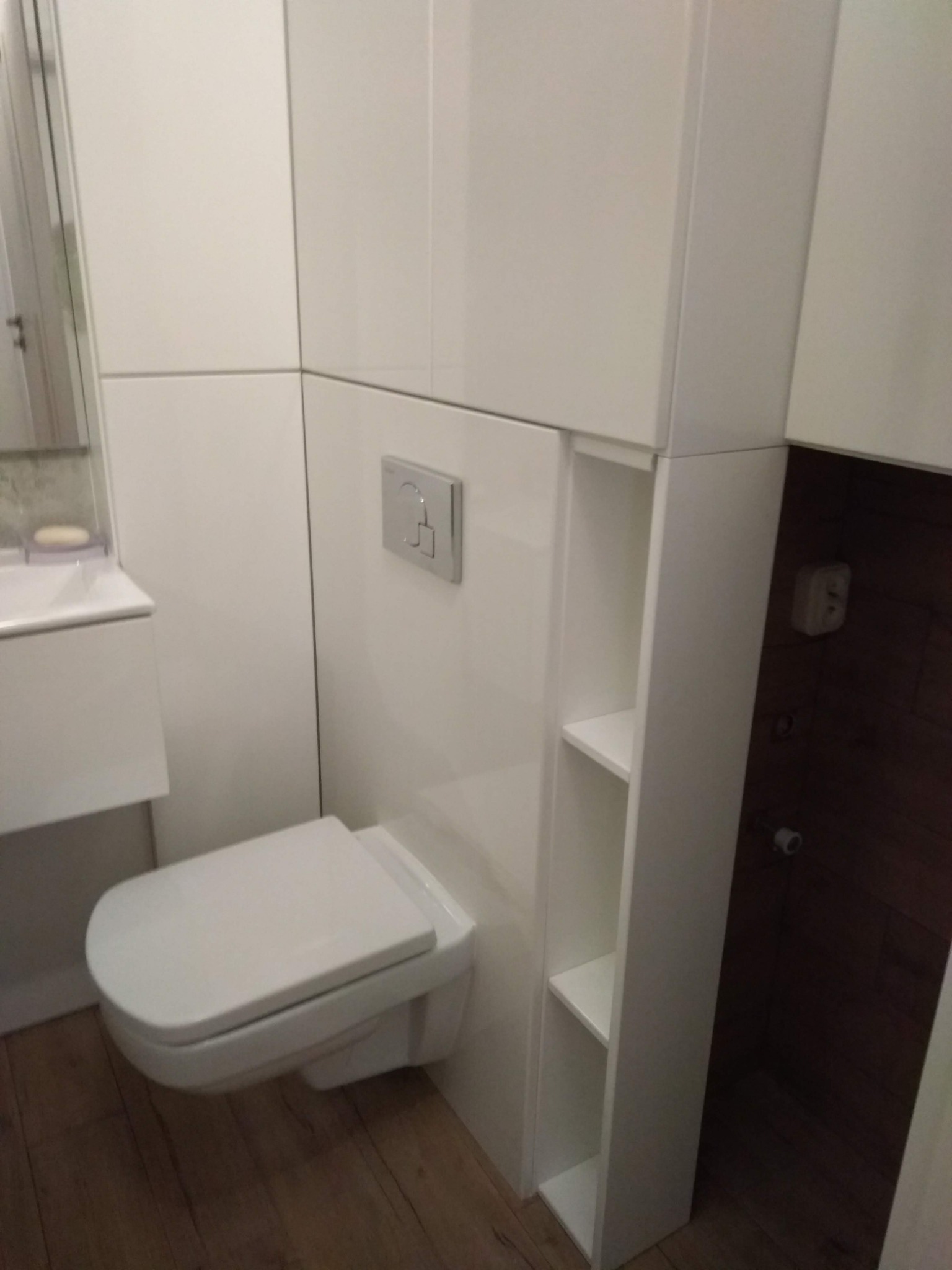 Nowoczesna, podwieszana toaleta w zabudowie meblowej z półkami w Golina. Białe, matowe fronty szafek, drewnopodobna podłoga i minimalistyczny design.