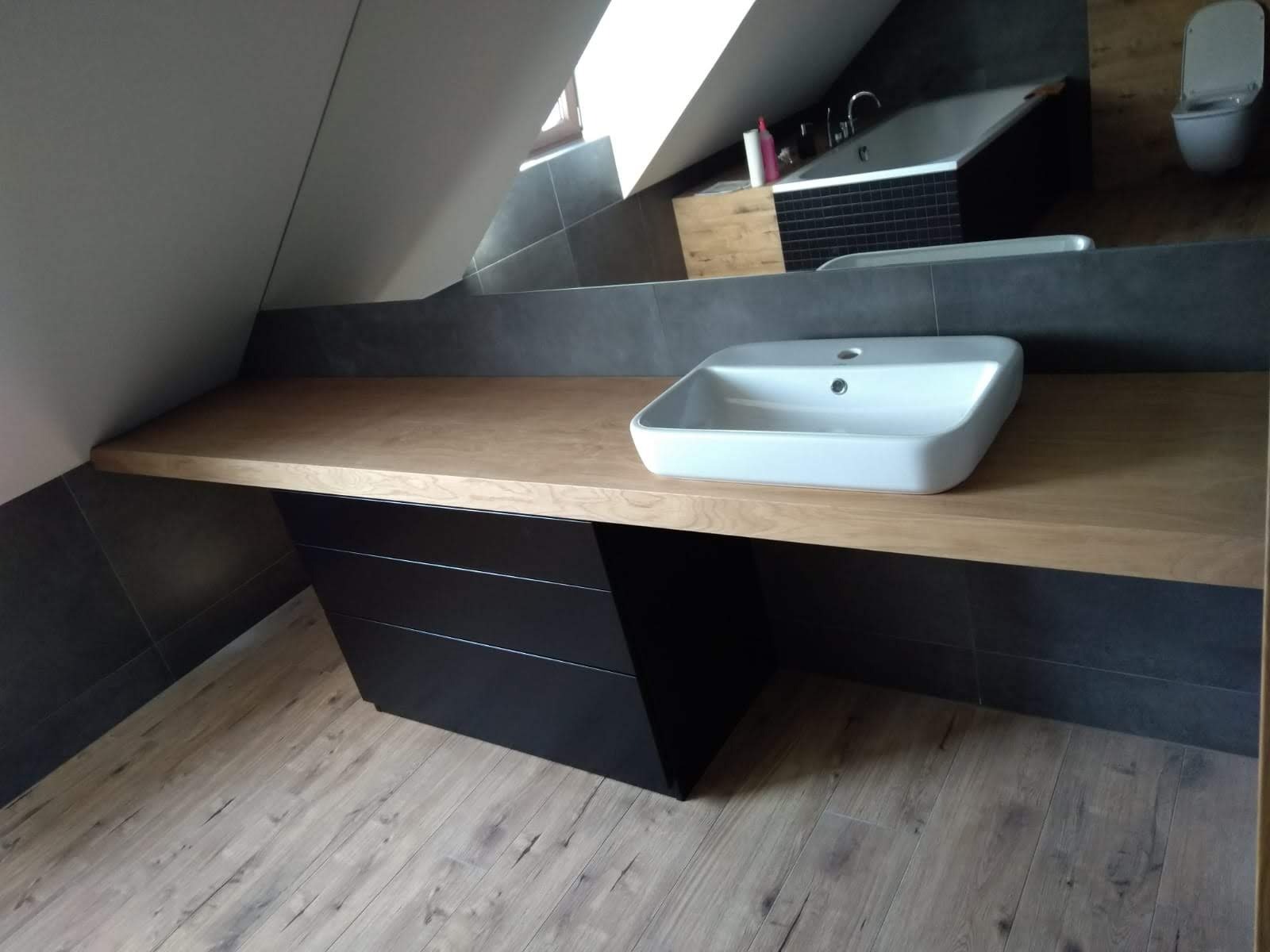 Łazienka na poddaszu z drewnianym blatem pod umywalkę, czarne szafki pod blatem, płytki imitujące drewno na podłodze, szare płytki na ścianach, wanna w tle.