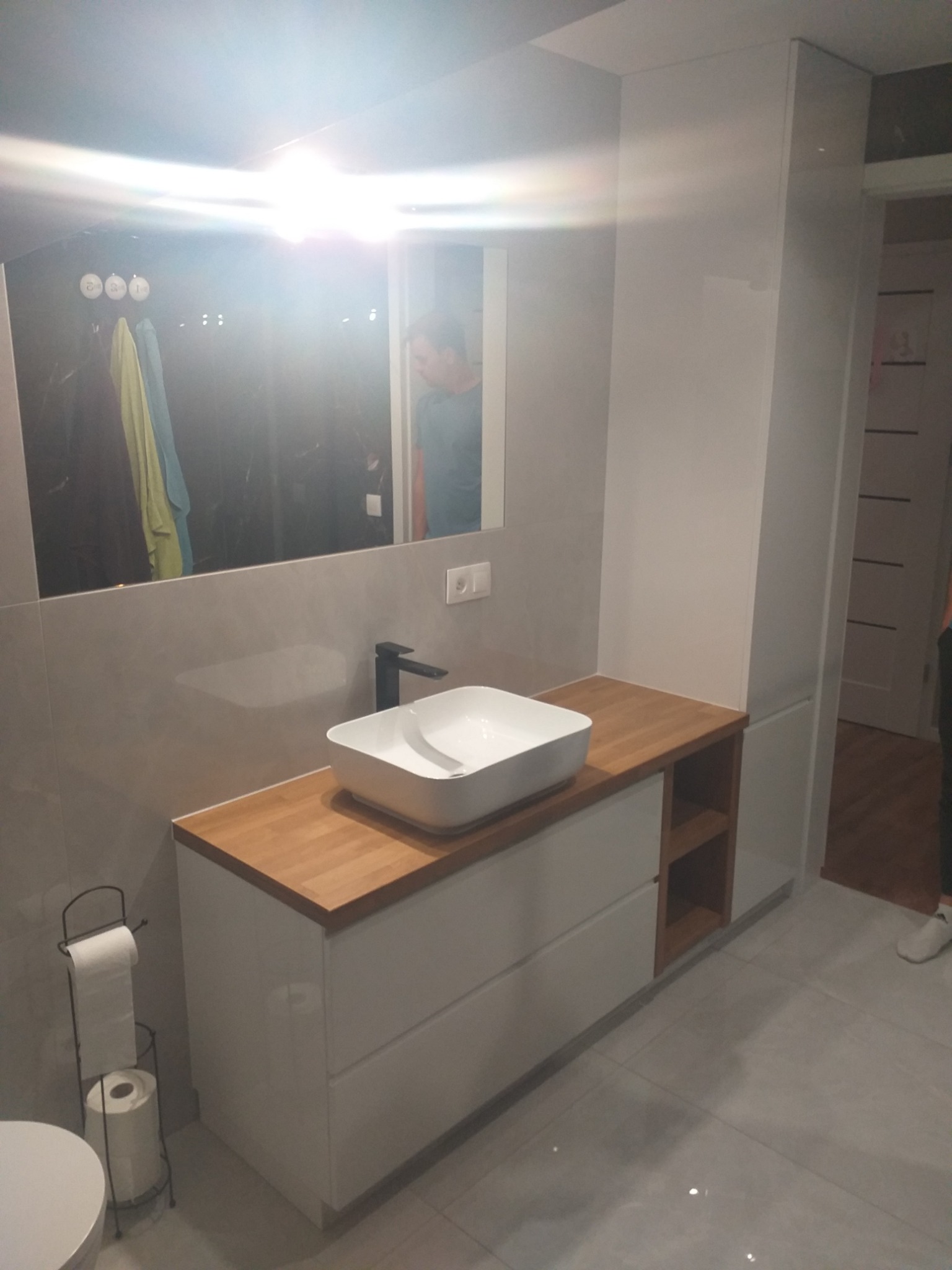 Nowoczesna łazienka z białą szafką pod umywalkę z drewnianym blatem, minimalistyczna armatura, lustro z widocznym odbiciem osoby, szara płytka podłogowa, widoczny fragment toalety i stojak...