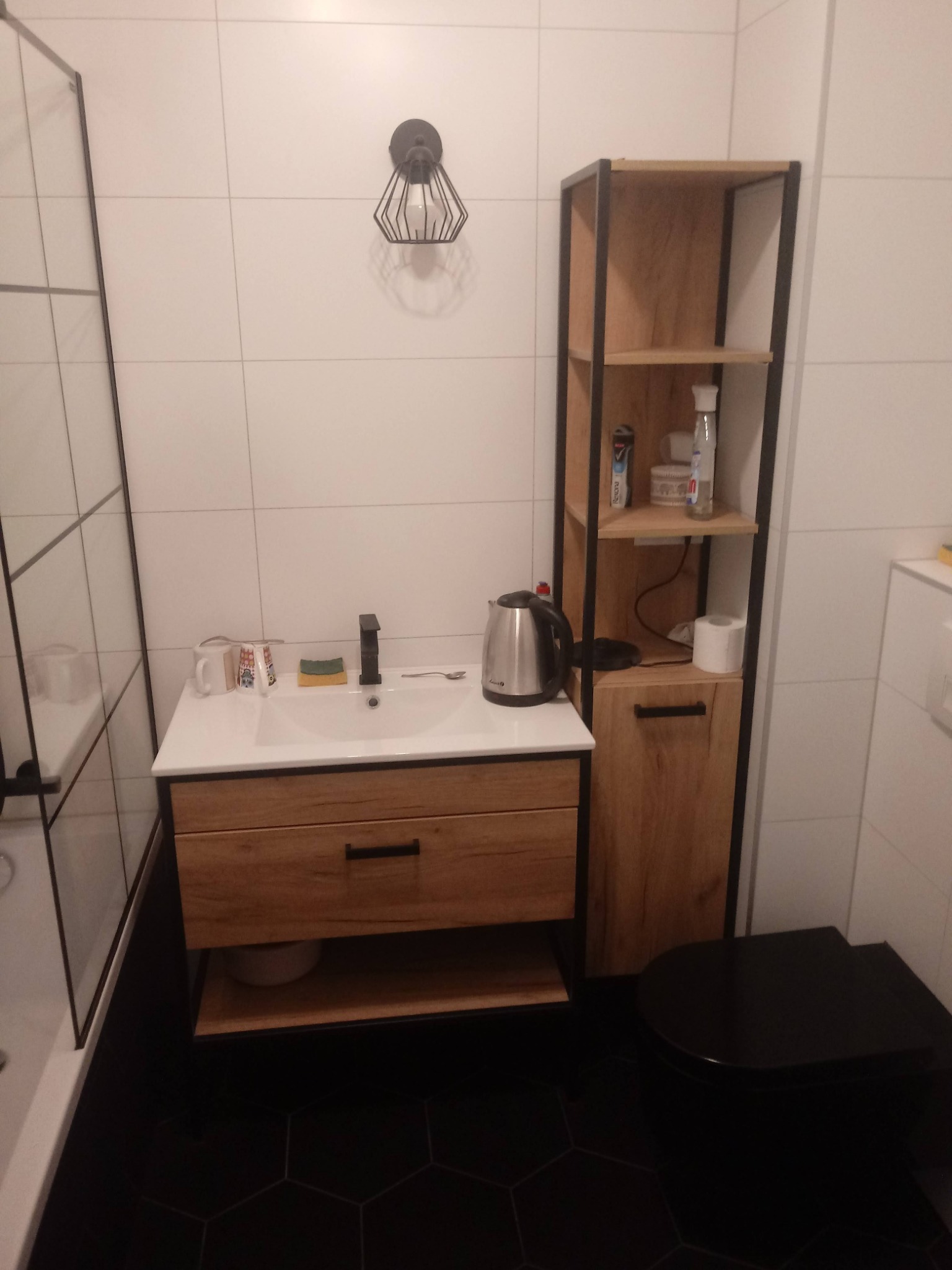 Nowoczesna łazienka z drewnianą szafką pod umywalką, czarną armaturą i heksagonalną podłogą, uzupełniona o metalowy regał z drewnianymi półkami i dekoracyjną lampę ścienną.